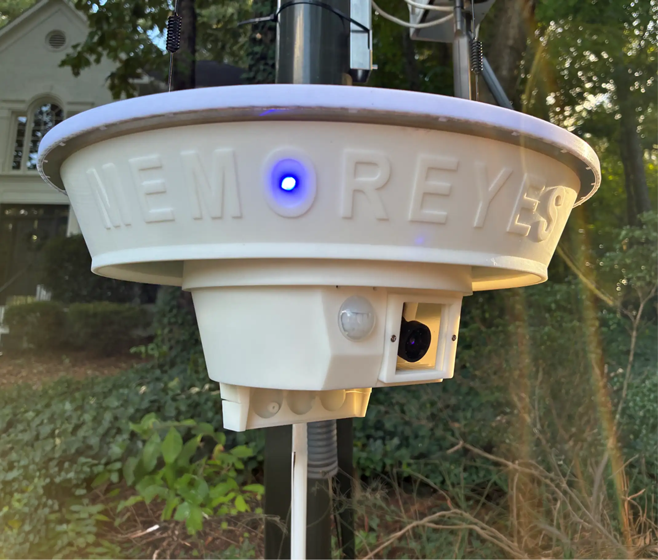 A white, round, outdoor security camera with a blue light on its front, mounted on a pole in front of a house with trees in